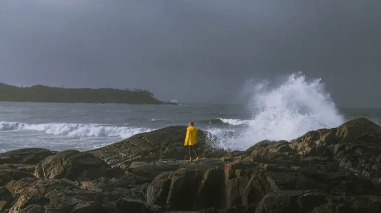 A woman in a yellow raincoat standing on the rocks by the waves in Tofino